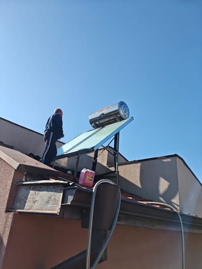 An outdoor, low-angle shot captures a man on a roof, likely inspecting or working on a solar water heating system under a clear blue sky. The scene is set in Kulata, Bulgaria.

The primary subject is a man, seen from behind, wearing a dark blue tracksuit with white stripes on his pants. He appears to be an older adult with short grey hair and is looking towards the solar water heater.

The most prominent object on the roof is a "Bartes" branded solar water heater. It features a shiny, cylindrical storage tank in silver, positioned above a large, angled flat-plate collector. The system is mounted on a dark metal frame. Near the solar heater, on the roof, there is a red plastic container with some unreadable text on its label, possibly containing liquid for maintenance. A white hose or pipe runs from the roof downwards, indicating connections to the building below.

The roof itself is composed of dark tiles and has exposed wooden or metal beams painted brown. The building below features textured, light brown stucco walls. Strong sunlight indicates a clear day, likely midday or early afternoon, with distinct shadows cast by the roof edges and the solar panel structure. The overall impression is one of maintenance or installation work being performed on the roof of a residential or commercial building.