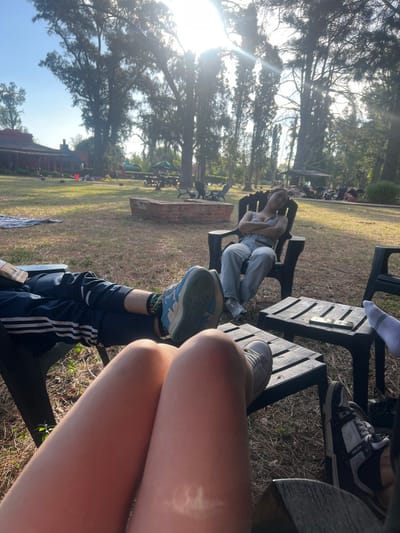 A group is relaxing outdoors on benches. One person in overalls reclines, facing away. Visible are legs and shoes of others. The scene features a grassy area with trees in the background. A brick fire pit stands nearby. The sun is shining brightly, causing lens flare.