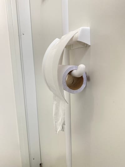 An extreme close-up shot depicts a partially used roll of toilet paper mounted on a broken, white plastic holder attached to a white, paneled wall. The toilet paper roll is nearly empty, with a cardboard tube visible. Some toilet paper hangs loosely from the roll. The holder is damaged, with pieces of the plastic broken off. The background is a white, paneled wall with visible screws. The location in Luzhitsy, Russia is unverifiable from the image.