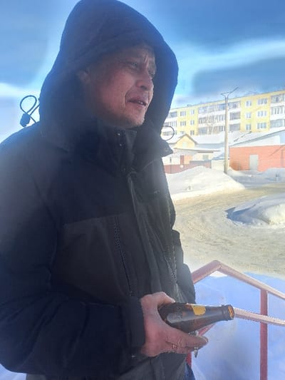 The image captures an adult male in a winter outdoor setting in Bolshaya Elhovka, Russia. The individual, seen in profile facing right, wears a dark, possibly black or navy, hooded winter jacket with the hood raised. A dark coiled wire object is visible behind his left ear/neck area. He exhibits stubble on his face and holds an amber-colored glass bottle, appearing partially empty with a yellow label on its neck, in his left hand. A ring is present on the fourth finger of his left hand. In the foreground, a reddish-orange metal railing is partially visible. The background features extensive snow cover across the ground. A prominent yellow multi-story apartment building with white window frames and balconies is situated behind smaller, single-story buildings, one of which is red brick. The sky above is largely clear and blue, with a faint horizontal cloud formation in the upper right. Utility poles are discernible in the mid-ground.