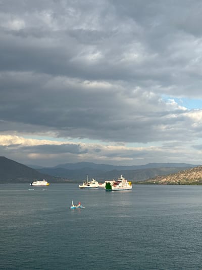 The image depicts a wide-angle view of a body of water with several ferries and a small boat. In the background, a chain of mountains borders the horizon, with dry, scrubby hills in the foreground. The sky is filled with dramatic, dark clouds, with patches of lighter sky and sunshine breaking through in the upper right quadrant.

The main subjects are three large ferries. One ferry, mostly white with a yellow stripe, is positioned on the left side of the image, sailing away from the viewer. In the center, a larger ferry, predominantly white with green and red accents, is also moving away. To the right, a third ferry, predominantly white with some red markings, is partially visible, also moving away.

In the foreground, a small, blue boat with a white canopy and a red flag is visible, carrying what appear to be two or three people. The water is a deep blue-green, with visible ripples suggesting a gentle breeze.

The overall scene is a maritime landscape, likely in a coastal area of Indonesia, given the mention of Upt Tambak Sari. The weather appears to be changeable, with stormy clouds contrasting with moments of light. The image conveys a sense of travel and transit on the water.