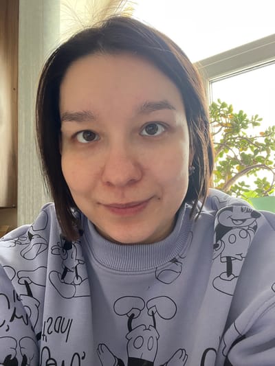 The image presents a close-up, head-and-shoulders portrait of a single female individual in an indoor setting within Kozlovka, Russia. The subject has short, dark brown hair, parted on the left side, and brown eyes. She displays a slight, closed-mouth smile and is looking directly into the camera. Her skin appears fair with visible pores and a subtle redness on the nose and cheeks. She is wearing a light lavender crewneck sweatshirt patterned with numerous black outline illustrations of Mickey Mouse characters. A small, dark-colored earring is visible in her right ear. The background on the left side of the frame shows a light brown, possibly wooden, surface. On the right, a window with a white frame reveals a bright, overcast sky and a green potted plant, likely a succulent, with numerous small, roundish leaves, positioned on what appears to be a windowsill.