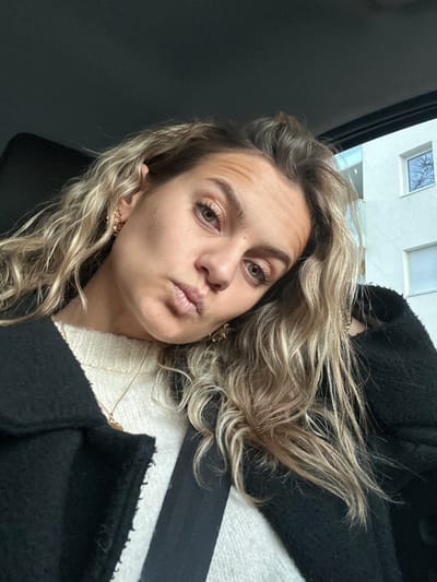 A woman angles her face toward the camera with pursed lips. Her wavy, blond hair is loose around her face and shoulders. She wears gold earrings and a white sweater beneath a black coat. A black seatbelt crosses her chest. A window showing a building with a partially visible tree is in the background. The setting appears to be inside a vehicle.
