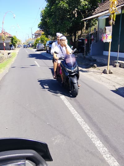 Two people are riding a black scooter down a paved road in a tropical setting, likely in Tabanan, Indonesia. The scooter has a blue tinted windshield with visible text that reads "DK 3458 OBO". The riders are dressed in white, with headbands also in white. The person in the back is holding a hand to their face, possibly indicating distress or blocking the sun. The scene is brightly lit, suggesting it is daytime with clear skies. Lush green trees line the road, and there are houses and other vehicles visible in the background, including several cars parked on the side of the road. Decorative Balinese banners, called "penjor", are seen hanging over the road and on the sides, which are typical during local ceremonies and festivals. The road has a white dashed line marking its center. The overall atmosphere appears to be a sunny day with ongoing local activity.