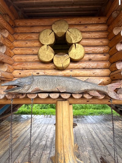 This is a full, eye-level, outdoor shot of a wooden structure in the city of Raifa, Russia. The structure is built from large, rounded logs, and features a decorative element above a carved wooden fish. The decorative element is a circle of six round, golden-colored logs, with a dark opening in the center. The carved fish is gray and textured to resemble scales, with its mouth open and its body positioned horizontally. It is suspended from the wooden structure by chains. Below the fish, there is a wooden floor, also constructed from planks. Two metal chains hang from the wooden structure, suggesting the presence of a swing, though no swing seat is visible in the frame. The background shows lush green grass and foliage, indicating an outdoor, possibly natural setting. The wooden surfaces appear weathered.