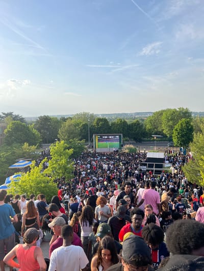 The image shows a large crowd of people gathered outdoors in Noisy-le-Grand, France, likely watching a sports game on a large screen. The sky is partly cloudy. The crowd appears dense, with people of diverse backgrounds. Trees are visible around the crowd. A large screen is showing a soccer game. The general atmosphere is one of excitement and public gathering.