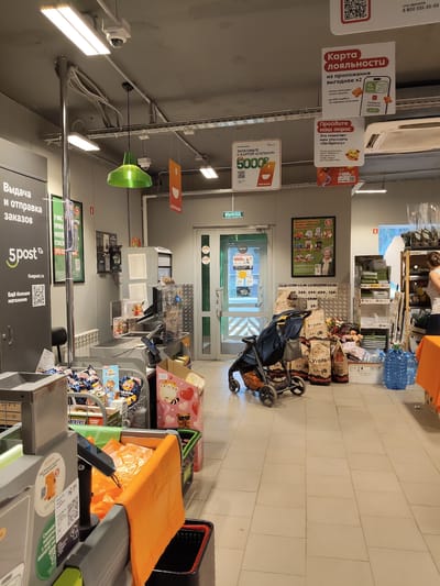 The image depicts the interior of a retail store, specifically a checkout area, likely in the city of Khodaevo, Russia. The scene is brightly lit with fluorescent lights and a prominent green pendant lamp. In the foreground, a gray checkout counter is visible, equipped with a scanner, a payment terminal, and an orange reusable shopping bag. Behind the counter, shelves are stocked with various products, including bags of charcoal, packaged snacks, and bottled water. A blue and black baby stroller is positioned to the left of the counter. In the background, a glass door with a "EXIT" sign is visible, leading to the outside. Several promotional posters are displayed on the walls and ceiling, advertising discounts, loyalty programs, and customer surveys. There are no visible people actively engaged in transactions, but a woman can be seen browsing shelves in the far right background. The overall environment suggests a typical supermarket or convenience store setting.