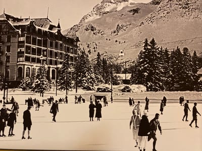 The image depicts a snowy, mountainous landscape with a large, multi-story building that appears to be a hotel or resort. In the foreground, a group of people are ice skating on a frozen surface. The individuals are dressed in vintage winter clothing, with some wearing skirts and dresses, and others in trousers and jackets. Several people are actively skating, while others are standing or walking. In the background, snow-covered evergreen trees line the area, and a ski lift cable is visible traversing the mountain. The overall scene suggests a winter recreational activity in a scenic, mountainous location. The provided information states the location is the city of Ленинское, Russia.