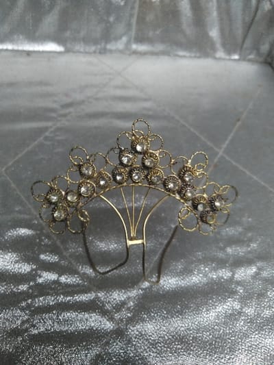 The image displays a decorative hair accessory, identified as a hair comb or small tiara, positioned in the city of Косшы, Kazakhstan. The object is crafted from a gold-toned metal, showing some signs of tarnish or patina. Its design features an upper arc composed of approximately fourteen interconnected floral motifs, each centered with a clear, faceted round gemstone, likely a rhinestone. These motifs are formed by delicate, looped metalwork. Below this decorative arc, a series of thin, fan-shaped metal wires converge to a central horizontal bar, from which two U-shaped prongs extend downwards. The accessory rests on a light grey or silver-colored fabric with a shimmering, possibly satin or crushed velvet, texture. No individuals are visible in the frame, and no actions are occurring.