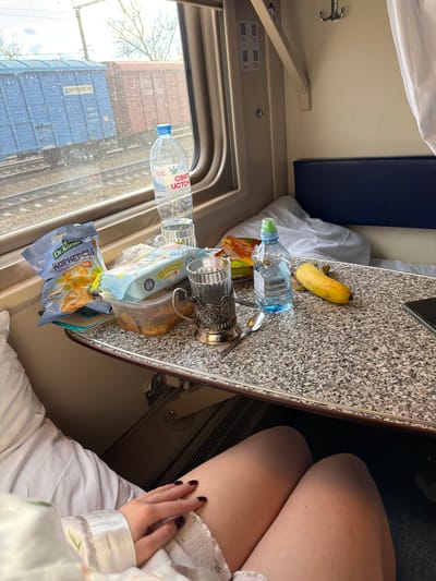 The scene is an interior view of a train compartment, likely taken from the perspective of a seated passenger. A table, covered in a speckled countertop, dominates the foreground and is laden with various items: a banana, a bottle of water, several packages of chips and snacks, a drinking glass in a metal holder with a spoon. The window to the left reveals a view of train tracks with cargo containers. A second bottle of water is visible through the window. Part of a blue cushioned seat can be seen in the background. A woman's thighs are also visible, suggesting she's seated near the table with one hand resting on her leg. The presence of these items suggests the passenger is on a journey.

I am unable to ascertain the exact location (Krasnyy Sulin, Russia) from the visual elements within the image. I am also unable to perform geolocation analysis using the image.