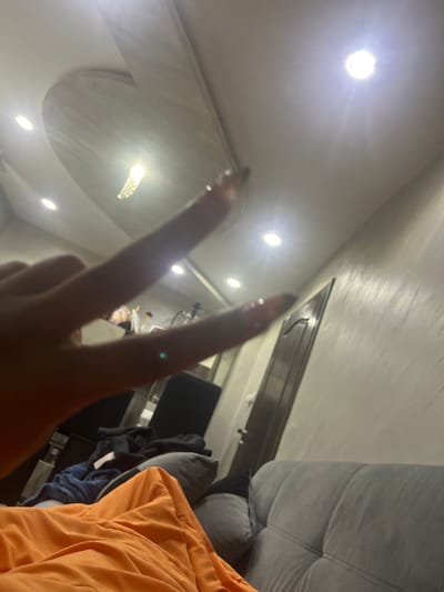 The image shows a low-angle view of a room with a ceiling featuring multiple recessed lights and a decorative, elliptical element. In the foreground, a hand is making a "peace" or "victory" sign. The background includes part of a gray upholstered sofa and an orange piece of clothing (possibly a hoodie). A television screen and assorted items on a shelf are also visible. The location, Voiniagovo, Bulgaria, is not explicitly discernible from the image. No people other than the arm, hand and fingers are visible. No specific actions other than the position of the hand are visible.