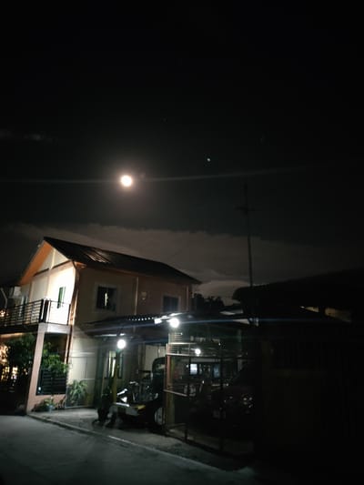 A low-angle, nighttime photograph captures a residential street in San Pascual, Philippines. A two-story house with an orange-trimmed roof and light-colored walls dominates the left side of the frame. It features a balcony with a dark railing on the second floor and several windows. Adjacent to the house, a covered parking area or carport is visible, with outdoor lighting illuminating the space. Inside the carport, a dark-colored vehicle and what appears to be a tricycle are parked. The foreground shows a paved road and sidewalk. The sky above is dark, with scattered clouds and a bright, full moon prominently displayed. The overall scene is dimly lit, with artificial lights from the house and street casting shadows.