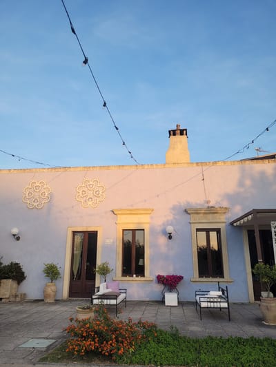 The image depicts the exterior of a charming building, likely a guesthouse or private residence, under a clear blue sky, bathed in the warm light of the golden hour. The building's facade is painted a soft periwinkle or dusty lavender hue, with the upper sections and a chimney exhibiting delicate pink and orange tones from the low-angle sunlight.

Three prominent windows and a door are visible, all featuring dark brown frames and ornate, light-colored stone or stucco surrounds. Two large, intricate white circular decorations, resembling lacework or mandalas, adorn the wall, casting soft shadows. Spherical outdoor light fixtures are mounted on the facade.

Across the upper left of the frame, string lights with bare bulbs are strung, hinting at evening ambiance. The foreground features a paved walkway made of grey rectangular stones. Along the right side of the path, a vibrant bush with numerous small orange flowers, possibly Lantana, adds a bright splash of color, complemented by lush green ground cover. Several potted plants, including small trees in terracotta planters, are placed around the outdoor area. Outdoor furniture includes a black metal bench with white cushions and a small white table displaying a pot of fuchsia flowers.

The scene suggests a peaceful, inviting atmosphere, characteristic of a pleasant early morning or late afternoon. A small rectangular sign near the upper right window reads "ALBA SALENTINA". Another partially visible sign below the middle window appears to indicate a street name, starting with "VIA DEI POETI...". The image is set in Sogliano Cavour, Italy.