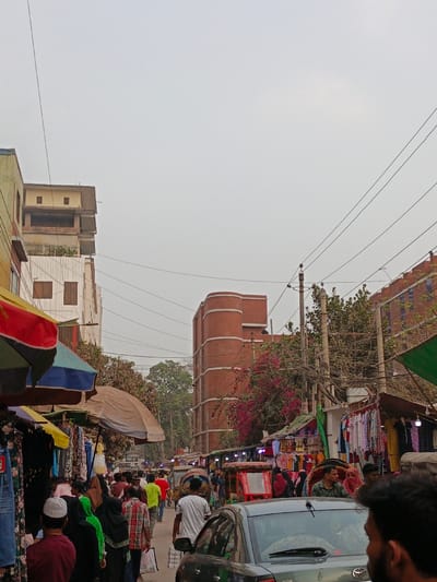 This image depicts a crowded street scene in Narayanganj, Bangladesh, under an overcast sky. In the foreground, the back of a dark grey Toyota car is visible, with a portion of a person's head and shoulder on the right, looking forward. To the left of the car, a dense group of pedestrians, including men, women, and children, walks along the street. Several individuals wear traditional attire, such as a white skullcap and a green hijab.

Along both sides of the street, makeshift market stalls are set up under colorful awnings and umbrellas (red, blue, brown, green), displaying various clothes, including dresses and shirts hanging on racks. Numerous electrical wires crisscross overhead, connected to a utility pole on the right side of the street.

In the mid-ground, beyond the immediate crowd, more people are visible, along with at least two cycle-rickshaws, one red and one black, suggesting ongoing traffic. A red brick building with distinctive rounded corners stands prominently on the right side, partially obscured by trees with pink blossoms and dense foliage. Another multi-story building, lighter in color with exposed brick on upper levels and visible window frames, is situated on the left side, behind a yellow-painted structure. The overall environment suggests a vibrant, bustling urban market area.