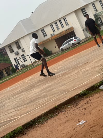 In Amansee, Nigeria, two young men are captured on a sports court. One, wearing a white t-shirt and black shorts, is in mid-stride, dribbling a basketball. His companion, dressed in a black t-shirt and black shorts, stands nearby, facing away from the camera. Behind them, a light-colored, multi-story building with a white roof and numerous windows stands prominently. A silver van is parked in front of the building. The court itself is a mixture of concrete and reddish-brown dirt. In the foreground, the ground transitions from the court to a dirt path, with some grass and scattered litter visible.