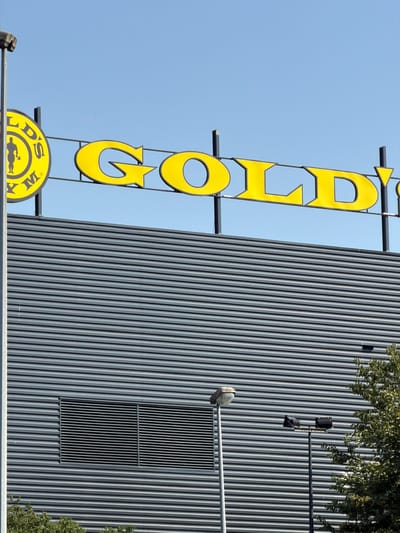This media file shows a close-up view of a vertical, yellow sign with the text "GOLD'S GYM" written on it. The sign is illuminated and appears to be made of metal. The letters are large and bold, with a black outline. The sign is mounted on a dark gray, corrugated metal building. The building has a series of vertical lines, giving it a ribbed appearance. There is a vent on the building, as well as some trees and branches visible at the top and to the right. The sky is a clear, bright blue, suggesting it is daytime and the weather is fair. The image appears to have been taken from a low angle, looking up at the sign and building. There are no people visible in the media file. The overall impression is of a commercial establishment. The text "GOLD'S GYM" is clearly legible, indicating the name of the business. The location context provided is Rungis, France.