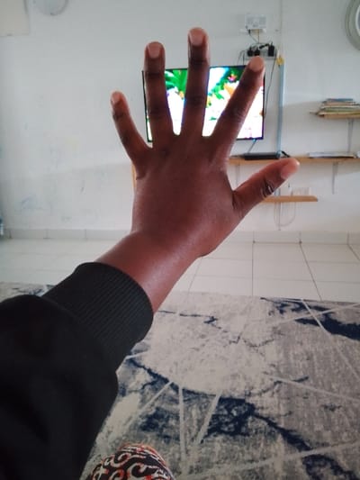 The image depicts an indoor scene in Olkeri ward, Kenya, dominated by a human left forearm and open hand in the foreground. The hand, with a dark skin tone, is held upright, palm facing away from the viewer, with all five fingers extended and spread. The forearm is covered by a black long-sleeved garment featuring a ribbed cuff. In the background, a flat-screen television is mounted on a white wall, displaying colorful, possibly animated, content. Above the television, a white electrical outlet or power strip with connected cables is visible. To the right of the television, two light-colored wooden floating shelves are affixed to the wall; the upper shelf holds a stack of multiple books or papers. The wall has faint blue smudges on the left side. The floor is covered with light-colored square tiles, partially obscured by a patterned rug featuring abstract designs in shades of grey, white, and dark blue. A small portion of patterned fabric, possibly clothing, is visible at the bottom of the frame below the forearm.