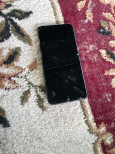 A black smartphone with a cracked screen lies on a patterned rug. The rug has a cream-colored base with a repeating floral pattern in shades of red, green, and brown. The smartphone is positioned vertically on the rug, with the top of the screen oriented towards the top of the image. The screen is dark and reflective, showing some smudges and the visible cracks. The overall scene suggests an indoor setting, possibly a home. There is no visible indication of the specific location within Kubanskaya, Russia.