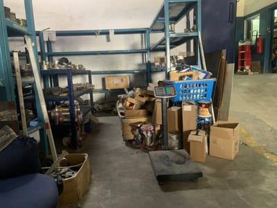 This image, recorded in Babawa, Nigeria, depicts the interior of an industrial storage or workshop space. The concrete floor features yellow demarcation lines on the right side. Blue metal shelving units line the white walls, holding various metallic components, including what appear to be pipe fittings or machinery parts, and numerous cardboard boxes. In the immediate left foreground, a dark blue fabric-covered object is partially visible next to an open cardboard box containing metallic items. The central floor area contains a digital industrial weighing scale, consisting of a display unit on a pedestal and a flat platform base. This scale is surrounded by numerous cardboard boxes, some stacked and open, containing diverse materials. A blue plastic laundry basket, heavily laden with items, rests atop a stack of boxes. Several cans, including a yellow and black paint or chemical container, are also present. Flat rectangular panels, possibly metal or wood, lean against a dark blue wall section on the right. An open doorway in the far right background reveals an adjacent room with additional shelving and a red wall-mounted fire extinguisher. No personnel are depicted within the scene.