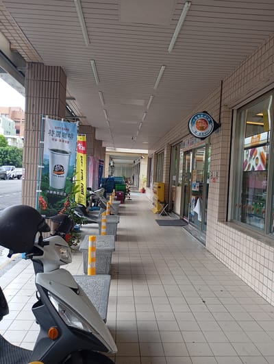 The image shows a covered walkway in front of a row of stores in Kaohsiung, Taiwan. In the foreground, there is a parked scooter with a black helmet. Along the walkway are several parked scooters interspersed with short, concrete benches featuring orange and white striped bollards. Banners advertising "CITY CAFE" are displayed next to the first store. Further down the walkway is a store with a sign for "We Are Family" ATM and a window displaying images of food. The walkway is paved with square tiles, and the building has beige tile walls. The ceiling is a light gray color with rows of fluorescent lights. Trees and buildings are visible across the street.