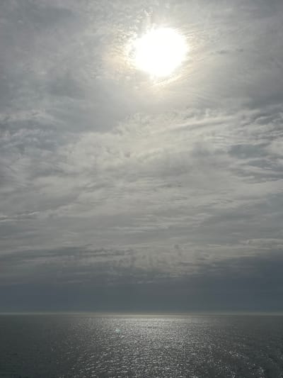A landscape photo showing the sea and a cloudy sky. The sun is shining brightly through the clouds, creating a shimmering reflection on the water. The location is unknown, France.