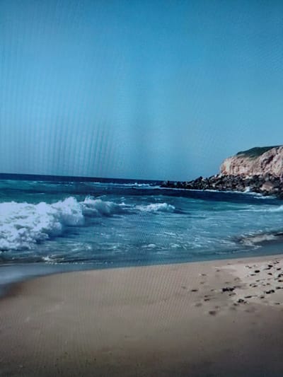 The image shows a beach scene with a clear blue sky and the ocean. The water is a deep blue and there are waves breaking on the shore. The beach is made of sand and there are some rocks and cliffs in the background. There are no people in the image. The image is a photograph of a natural landscape.