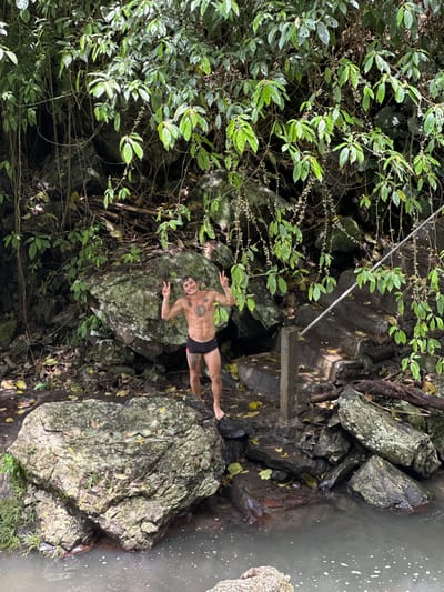 An adult male is observed standing barefoot on a wet, rocky riverbank in Ambengan, Indonesia. The individual is barechested, displaying a muscular physique and multiple tattoos across his chest, abdomen, and left thigh. He wears black swim briefs with a thin pink stripe along the side. Both of his arms are raised, with hands open, extending the index and middle fingers in a "peace" gesture. He is smiling towards the viewer. Behind the subject, the environment is characterized by large boulders, damp earth, and dense green tropical foliage, including numerous hanging vines. To the subject's immediate right, a concrete stairway equipped with a metal handrail descends towards the water. A body of murky water with minor surface agitation is visible in the lower foreground and extending to the mid-right of the frame.