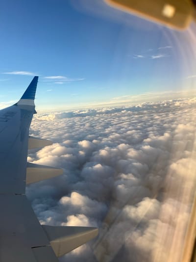 The image presents an aerial view captured from the window of an aircraft. The aircraft's left wing and its winglet are visible in the lower-left portion of the frame, featuring a light blue and white striped livery and the partial registration "LV-KHP" on its surface. Below the aircraft, a vast, unbroken expanse of white, cumuliform clouds extends to the horizon, illuminated by soft, warm light, suggesting either sunrise or sunset. Above the cloud layer, the sky is clear blue, gradually deepening in hue towards the top of the frame. A blurred reflection of an interior aircraft component, possibly an overhead panel, is visible in the upper-right corner of the window. No ground features or specific geographical landmarks are discernible, making it impossible to ascertain the location as Colonia del Sacramento, Uruguay, as the scene is entirely above a dense cloud cover.