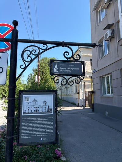 This is a photograph taken in Taganrog, Russia, depicting a street scene with informational signs. The image is oriented vertically. In the foreground, a black wrought-iron pole holds two signs. The upper sign, also black, has white text in Cyrillic that translates to "Greek Male Monastery was located here" and displays an icon of a church. Below it, a larger black sign has a black and white illustration of a church and a QR code. The text on this larger sign, also in Cyrillic, provides a historical description of the monastery's founding in 1813, its patrons, architectural style, and eventual closure and demolition in the 1920s and 1930s. A red and blue traffic sign is also visible on the pole.
In the background, a modern building with air conditioning units is visible, suggesting an urban setting. Sunlight indicates it is daytime, with clear blue skies. The overall scene captures a blend of historical information and contemporary urban life, marking a significant historical site within the city.