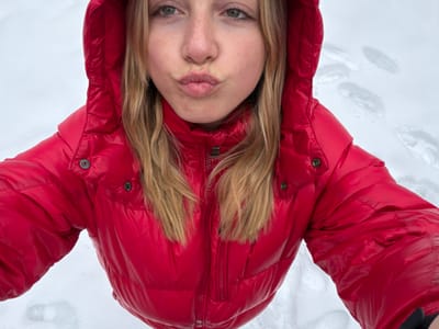 A close-up, high-angle selfie depicts a single individual, likely female, with light blonde hair, positioned against a snow-covered background. The individual is wearing a bright red, shiny, hooded puffer jacket, zipped up to the neck. Their face is partially visible, showing light-colored eyes and scattered freckles across the nose and cheeks. The lips are pursed in a distinct "kissing" expression, directed towards the camera. In the background, to the right of the subject's head, multiple indistinct footprints are pressed into the white snow. The scene is located in La Clusaz, France.