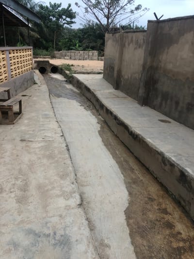 The image showcases an exterior concrete structure with visible weathering and aging. A large overhanging concrete element casts shadows on the lower structure. The concrete ground has variations in color, possibly due to moisture or wear. Two dark drainage pipes are visible along the structure. A partial view of a building and trees is in the background.