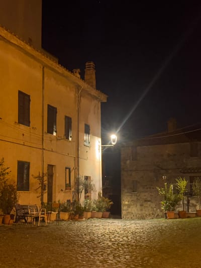 The image depicts a night scene in Castelnuovo di Porto, Italy. The view is angled upwards towards a multi-story building with closed window shutters. The building is illuminated by a street lamp. Adjacent to the building is a stone wall. A cobblestone pathway runs alongside the wall. Potted plants are situated along the base of the building and wall. There are some chairs and a table on the right side of the image. The sky is dark.