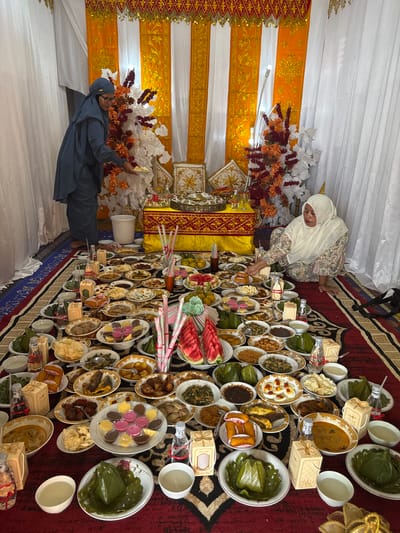 In a room decorated with orange and white drapes, a feast is laid out on the floor. Two women in Islamic attire are present. The woman on the left, wearing a blue hijab and glasses, is holding a small plate of food and appears to be serving. The woman on the right, in a cream-colored hijab and patterned dress, is seated and reaching towards the food. The floor is covered with a red, patterned rug. The spread includes numerous dishes of food, rice wrapped in banana leaves, fruit, drinks, and decorative boxes. The scene suggests a cultural or religious gathering, likely a celebration or offering. The location is specified as Kruengseupeng, Indonesia.