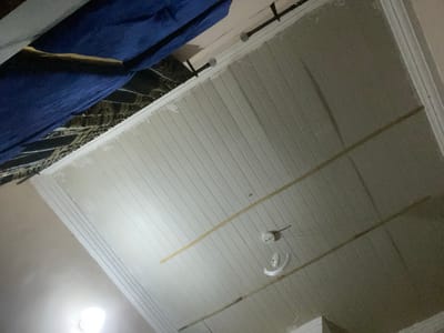 The image depicts a ceiling with white, grooved paneling. There are two thin, wooden slats running horizontally across the panels. A smoke detector is mounted on the ceiling, with a white, curved object, possibly a decorative light fixture or part of a fan, hanging below it. In the upper left portion of the image, a dark blue fabric is visible, draped over something that appears to be a mattress or bed. The overall lighting suggests it is nighttime or an interior setting with artificial light. The scene is likely a room in a residential or commercial building. There is no visible text in the image. The location context is Gbaza, Nigeria, but this cannot be determined from the image itself.