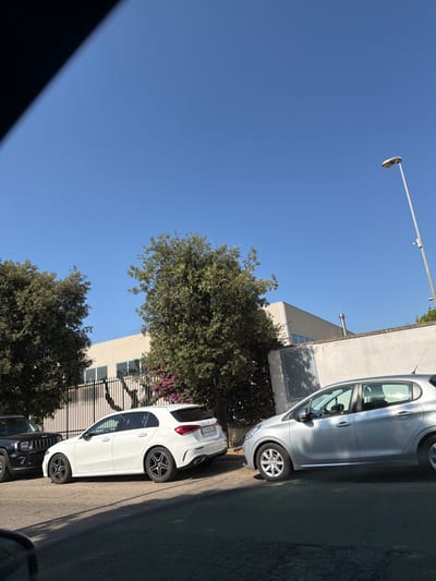 In the city of Cavallino, Italy, on a clear, sunny day, several parked vehicles are visible. In the foreground, a silver Peugeot 208 is parked on the right, facing left. To its left, a white Mercedes-Benz A-Class is parked. Further left, a black Jeep is partially visible. Behind the cars, a modern, light-colored building with large windows stands. A black metal fence separates the building from the street, and lush green trees with pink flowering bushes line the fence. A tall street lamp is positioned on the right side of the image, and a security camera is mounted on its pole. The scene is set on an asphalt road with visible cracks. The sky above is a bright, clear blue.