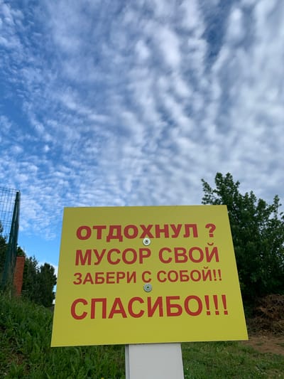 The image shows a yellow sign with red Russian text placed outdoors. The sign is mounted on a white post. The text on the sign reads: "ОТДОХНУЛ? МУСОР СВОЙ ЗАБЕРИ С СОБОЙ!! СПАСИБО!!!" which translates to "Resting? Take your trash with you!! Thank you!!!". The sign is positioned in front of lush green grass and vegetation. In the background, there is a tall fence, trees, and a partly cloudy blue sky. The image appears to be taken from a low angle looking up. The specific location in the city of Chernovo, Russia is not discernible from the visual information present in the image.