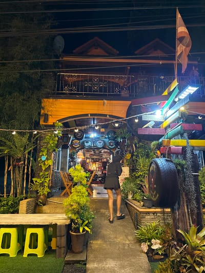 The image shows a nighttime view of a quirky, decorated outdoor establishment in Kalai, Thailand. A person, dressed in a dark t-shirt and shorts, stands facing away from the camera, looking into the open doorway of the establishment. The entrance is adorned with various decorations, including what appear to be car tires, plants, and colorful wooden beams. The establishment itself seems to be a bar or restaurant, as suggested by a sign that reads "BAR & RESTAURANT" with an eagle graphic.

The scene is lit by a mix of artificial lights from the establishment and string lights decorating the exterior. The overall atmosphere appears casual and inviting, with a collection of round decorative items, possibly clocks or signs, mounted on the wall inside. The ground is paved with concrete, and there are potted plants and bamboo structures around the entrance, suggesting a tropical setting. Yellow plastic stools are visible on the right side of the frame, placed on a patch of artificial grass, further adding to the eclectic decor. The sky is dark, indicating it is nighttime.