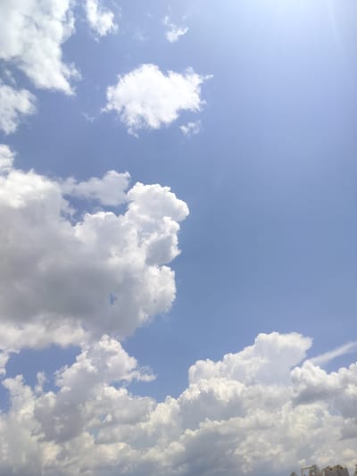 The image shows a bright blue sky filled with scattered white clouds. Some clouds are large and puffy, while others are thinner and wispy. Sunlight streams in from the top right of the frame, creating a slight lens flare effect. The bottom edge of the image shows the tops of some buildings and structures, suggesting an urban or industrial setting. The scene is a typical daytime sky, likely taken outdoors.