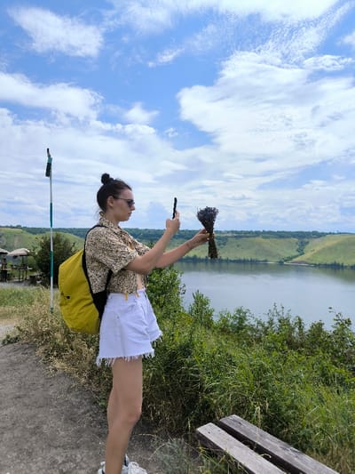 The image depicts an outdoor scenic view near Horaivka, Ukraine, overlooking a wide body of water, likely the Dniester River, with distant hills under a partly cloudy sky. A female subject stands in profile, positioned in the left-center of the frame, facing right. She wears a light-colored patterned short-sleeved shirt, white frayed denim shorts, and light-colored sneakers, with a yellow backpack on her back. Sunglasses are worn on her face, and her dark hair is pulled into a bun. Both hands are raised, with a black smartphone held in her left hand and a small, dark purple bundle of dried flowers, resembling lavender, held in her right hand, as if being photographed.
The immediate foreground consists of a dirt path with sparse dry grass. In the lower right corner, two parallel wooden planks of a bench are partially visible. To the left of the subject, a tall, slender pole with alternating green and white sections and a dark flag attached near its top is present. Beyond the subject, green shrubs and dense vegetation line the edge of the elevated ground, which slopes down to the calm, reflective water. Across the water, a panorama of undulating green hills, some with tree lines, extends into the distance. Faintly visible on the far left mid-ground, on the elevated terrain, are several outdoor tables shaded by circular umbrellas. The sky above is a light blue with scattered white and grey clouds.