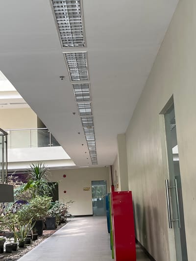 The image depicts an indoor hallway or atrium with a tiled floor and a white ceiling. Along the ceiling, multiple fluorescent light fixtures are visible in a linear arrangement. The hallway walls are painted a light, muted color. On the left side of the hallway, there is a prominent display of potted plants, including a tall, spiky plant and several smaller plants. To the right of the plants, a walkway extends into the distance, bordered by a wall on the right. In the distance, several colorful, rectangular cabinets are present, along with a glass-paneled door. A sign with the words "SERVICE ROOM" is affixed to the wall near the door. This scene is likely within a public building or commercial space in Koronadal City, Philippines.