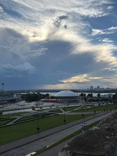 This is a high-angle, vertically oriented photograph looking down and across a landscape with a dramatic sky. The sky is filled with a mix of dark, heavy clouds and bright, sunlit clouds, suggesting dynamic weather. Sunlight breaks through in a few areas, creating a luminous effect.

Below the sky, a city skyline is visible across a body of water, with buildings of varying heights and styles. In the foreground, a large, domed structure, possibly a stadium or convention center, is prominent. Adjacent to this structure is a wide, paved area with multiple lanes and pedestrian walkways, bordered by neatly manicured green lawns. Several lampposts are visible along the walkways.

A stone wall forms the right edge of the frame, overlooking the scene. On the paved area, a few cars are present, and several people are seen walking along the pathways. One pair of individuals, dressed in a white top and blue pants, is standing near the stone wall. Another person in a blue shirt is walking along the pathway.

The lighting suggests it is either early morning or late afternoon, with the sun low on the horizon. The presence of both dark and bright clouds indicates varied weather conditions, possibly a transition. The organized layout of the paved area, lawns, and modern architecture points to an urban or recreational setting. No visible text is discernable in the image.