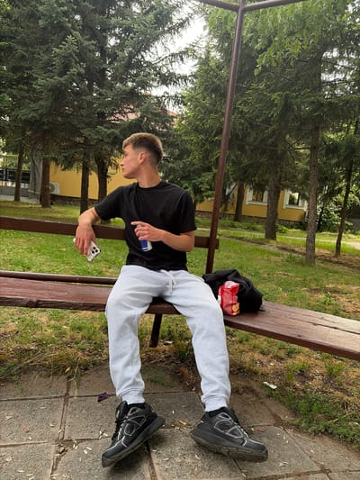 A young man is captured resting on a wooden park bench in an outdoor setting, likely in Slivnitsa, Bulgaria. He is seated with his legs stretched out, wearing a black short-sleeved t-shirt, light grey sweatpants, and black sneakers with white accents. His short, light brown hair is visible, and he is looking down, focused on a white smartphone (appears to be an iPhone) held in his left hand. In his right hand, he holds a blue and silver beverage can, possibly an energy drink, though the brand is not fully legible.
Next to him on the bench, there is a red bag of snacks, likely crisps or chips, and a folded black hooded garment. The bench itself is made of dark stained wood with metal supports.
The background is dominated by lush green foliage, primarily tall evergreen trees, suggesting a park or forested area. Beyond the trees, hints of yellow buildings with white window frames can be seen. The ground around the bench is a mix of grass, dirt, and paved grey flagstones. A dark brown metal bar runs horizontally across the scene in the mid-ground. The lighting suggests a bright, possibly overcast, daytime. The man's posture and focus on his phone indicate a moment of leisure or casual interaction. No other individuals are visible. No distinct legible text is present in the image.
