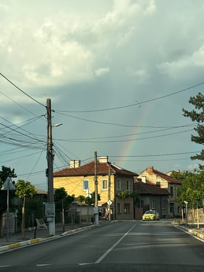 The image shows a street scene in Dolna Banya, Bulgaria, with a prominent rainbow arching across a cloudy sky. In the foreground, a paved road runs through a residential area. Utility poles with numerous tangled wires are visible along the roadside, along with streetlights. Several houses with red-tiled roofs are situated on either side of the street. One house is painted yellow, and another is a muted gray. A bright yellow car is parked on the right side of the road. Trees line the sidewalks, and there are some pedestrians in the background. The overall atmosphere suggests a post-rain or overcast day, with the rainbow indicating a break in the weather.