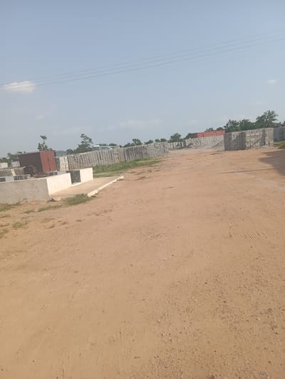 This image depicts an outdoor construction site in Tasha, Nigeria. The foreground is dominated by a dirt road with tire tracks visible. To the left, a low concrete structure with white and black decorative elements is present. Beyond this, a red shipping container is visible, partially obscured by stacks of what appear to be building materials. In the background, long rows of stacked concrete blocks stretch across the scene, with scattered trees and foliage interspersed. The sky is clear and blue with a few faint clouds and power lines running horizontally across the upper portion of the image.