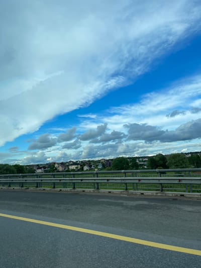 The image shows a roadside view from what appears to be a bridge or elevated roadway. A grey metal guardrail runs along the edge of the road. Beyond the guardrail, there is a line of buildings, including houses, partially obscured by trees. The sky above is blue with white and grey clouds. A yellow line marks the edge of the asphalt road. The picture's location is possibly near Бавыкино, Russia, due to the presence of buildings characteristic of Russian suburban or rural areas. The overall scene is calm, depicting a typical roadside view on a cloudy day.