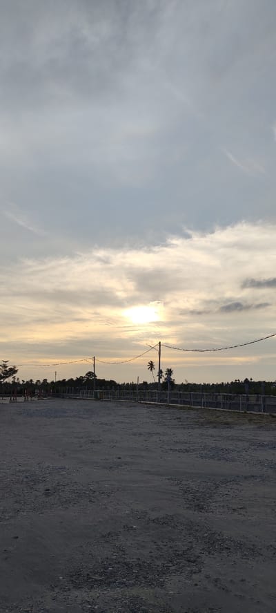 The image depicts an outdoor scene with a gravelly ground in the foreground, leading to a fence or barrier in the mid-ground. Beyond the barrier, there are scattered trees and vegetation, including palm trees. The sky is overcast with clouds, but a bright sun is visible through a gap, suggesting either sunrise or sunset. There are power lines stretching across the scene, supported by utility poles. The overall atmosphere is calm and serene. The location is identified as Ranto, Indonesia. No people or specific activities are clearly visible, making it a landscape or environment shot. The time of day is likely early morning or late afternoon due to the lighting conditions.