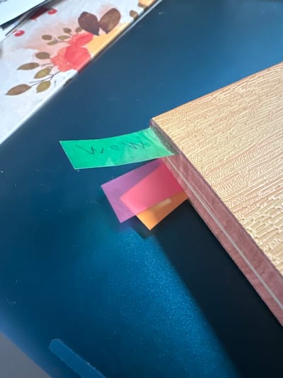 The image shows a close-up of a book with a textured, light brown cover. Several colorful sticky notes are protruding from between the pages. One green sticky note has handwritten text on it. Below the green note are a translucent pink note and an orange note. The book is resting on a dark blue, textured surface. In the upper left corner, a portion of a surface with a floral pattern, featuring red and pink flowers and green leaves, is visible. There are no people or specific landmarks of Trieste, Italy visible in the image.