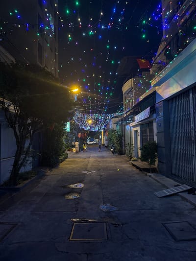 Captured at night in Nam Nha Trang, Vietnam, this image reveals a narrow street adorned with colorful string lights, which create a festive atmosphere overhead. A Vietnamese flag is visible amidst the lights. People can be seen walking in the distance, their forms illuminated by the strung lights and the nearby lamppost. The street itself is concrete with minor surface imperfections. Buildings line both sides of the street, their facades only partially visible due to the darkness. Some greenery is interspersed along the streetscape.