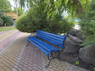 The image captures a vibrant blue park bench with ornate black metal legs, positioned on a decorative paved walkway. The walkway is composed of rectangular bricks in a repeating pattern of pink, beige, and gray. To the right of the bench, large, dark gray rocks are nestled amongst sparse greenery and fallen leaves. Behind the bench, a dense green shrub and the trunk of a mature tree with textured bark are visible, its branches draped with cascading green foliage. In the background, beyond the shrubbery, a large dump truck with a wooden bed is partially visible. The setting appears to be a park or a public outdoor space. The overall scene is tranquil and suggests a warm, possibly summer day. The location is identified as Kovalivka, Ukraine.