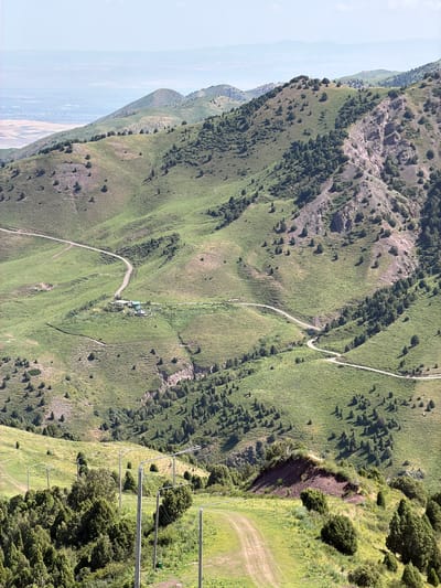 A wide shot captures a mountainous landscape in Kashka Suu, Kyrgyzstan, under a bright, hazy sky. Rolling green hills are dotted with scattered trees. A dirt road winds its way through the terrain, leading to a cluster of buildings at the base of a larger hill. The buildings appear to be a small settlement or farmstead. In the foreground, another dirt path is visible, leading up a grassy incline. The overall impression is one of a rural, natural environment.